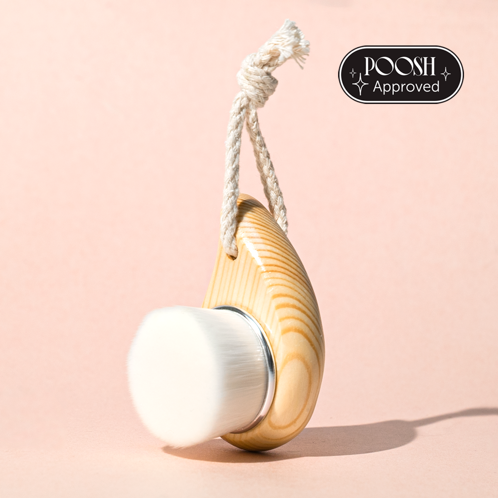 A facial dry brush with a wooden handle and synthetic bristles, hanging from a rope against a pink background.