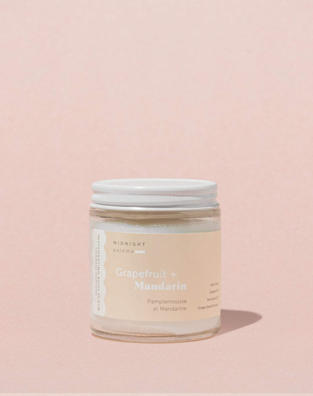 A glass jar containing Grapefruit + Mandarin Body Scrub, labeled with the product name and weight, placed against a pink background.