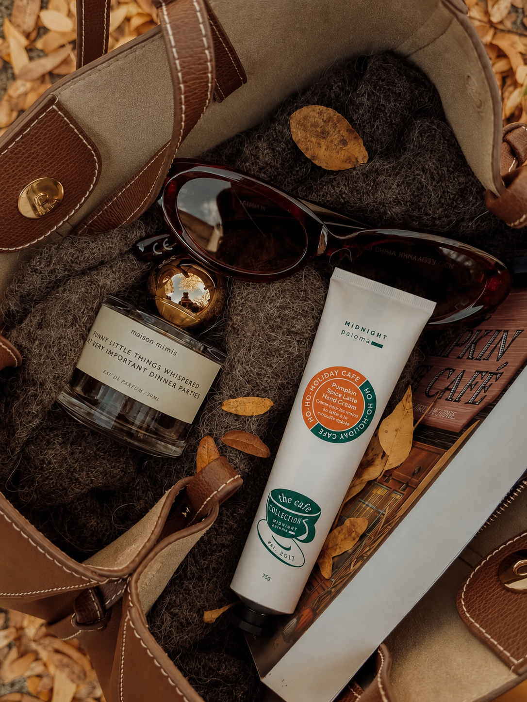 A styled flat-lay of a white tube of Midnight Radiance hand cream and a small glass jar of tea light candles on a dark gray blanket inside a brown leather bag, surrounded by autumn leaves.
