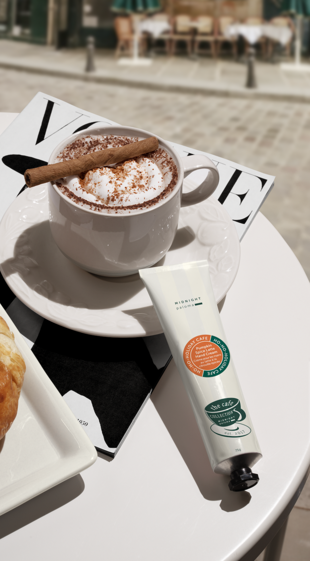 Hand cream tube next to a cup of hot chocolate on a magazine with a blurred outdoor background