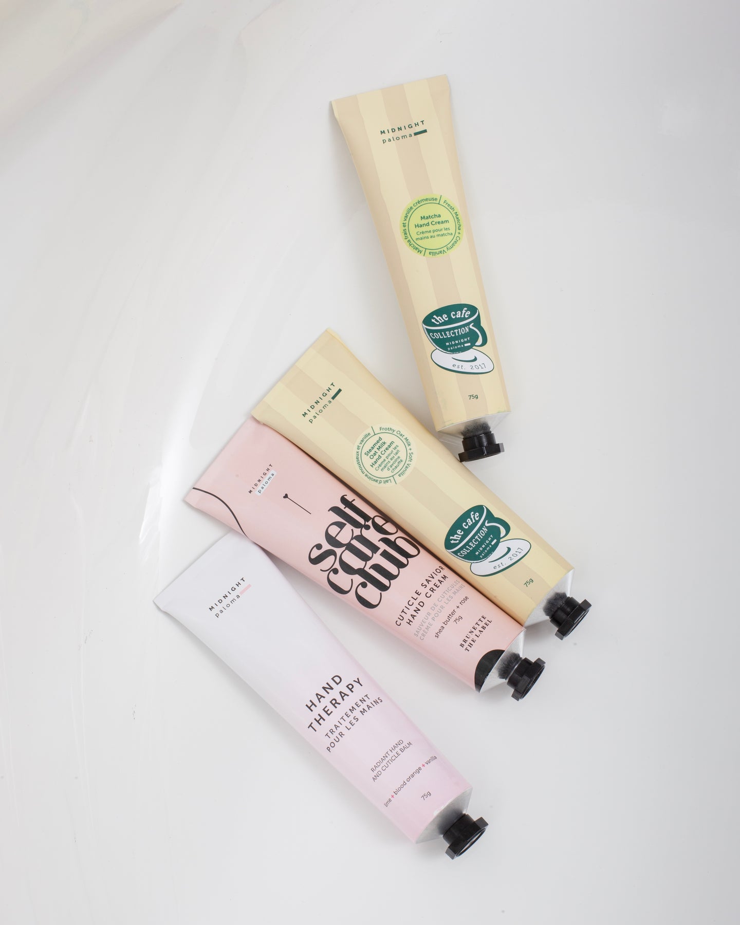 A tube of hand balm positioned next to its packaging, labeled 'Hand Therapy', with a color scheme of pink and white and an indication of blood orange, lime, vanilla, and grapefruit scents.