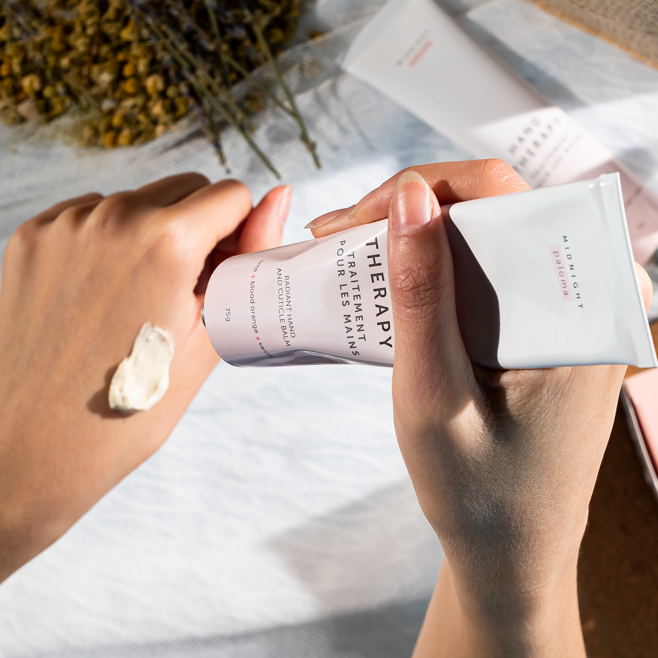 A tube of hand balm positioned next to its packaging, labeled 'Hand Therapy', with a color scheme of pink and white and an indication of blood orange, lime, vanilla, and grapefruit scents.